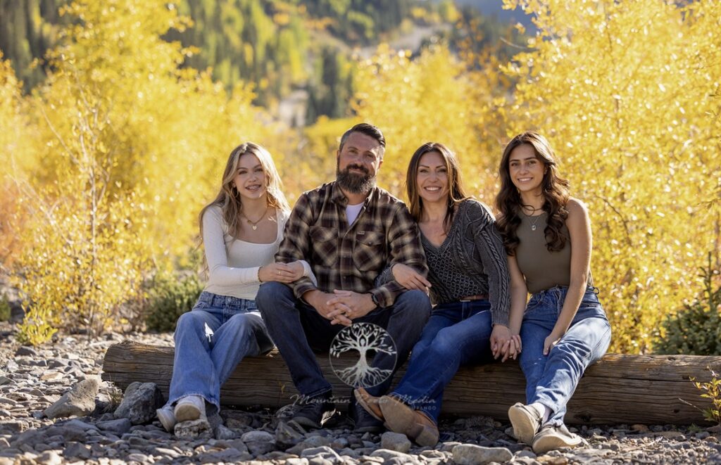 Jennifer Tewell and Family at Four Corners Child Advocacy Center A family of four, consisting of two adults and two young women, sitting on a wooden log amidst a scenic backdrop of golden autumn trees. All are smiling warmly.