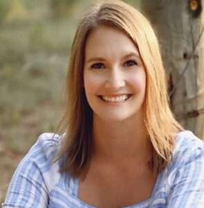 Portrait of a smiling woman with blonde hair, wearing a blue and white striped shirt, sitting outdoors against a tree.